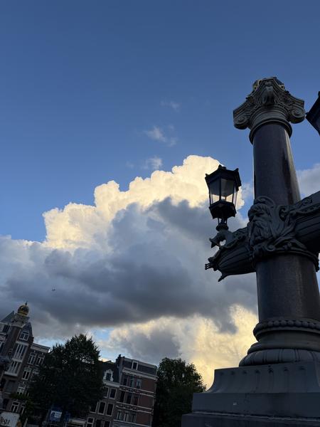 The photo captures a dramatic sky over Blauwbrug in Centrum, Amsterdam, featuring an ornate lamppost in the foreground